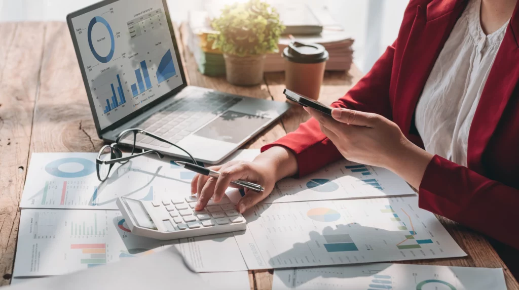 A person using a smartphone and calculator at a cluttered desk with charts and a laptop, representing CFO services and small business financial planning.
